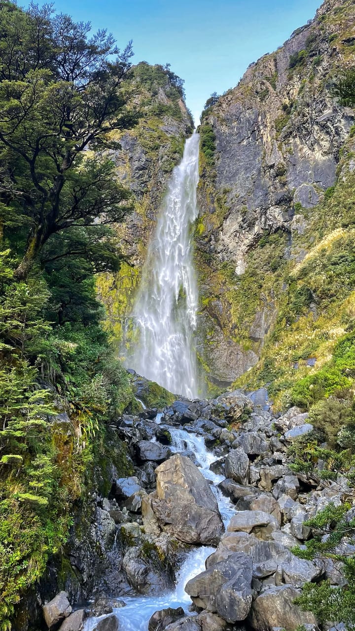 A ginormous waterfall cascades down a rock face, emerging into a rocky stream. Moss grows against the rock.