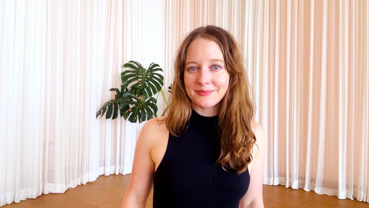 Clara Ritger looks directly at the camera in a black turtleneck. She is in a pale pink studio space with a green fern and wood floors.